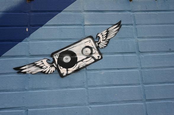 Graffiti of a black and white audio cassette with cartoon eyes and wings on both sides, painted on a blue brick wall.

Alt-Text (DE):
Graffiti eines schwarz-weißen Audiokassettenmotivs mit Comic-Augen und Flügeln auf beiden Seiten, gemalt auf eine blaue Ziegelwand.