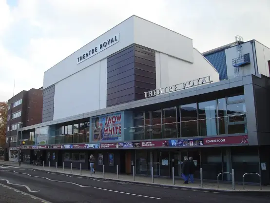 The image shows a building identified as "Theatre Royal." It appears to be a modern theater structure, featuring large windows and a marquee advertising a production, likely related to "Snow White." There are people walking in front of the theater, and the setting looks urban with other buildings nearby.

Image Credits: Wikimedia / Northmetpit / Public domain