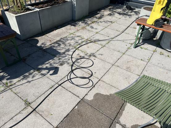 Here's an alt-text description for the image:

A sun-drenched patio with grey paving stones shows a green slatted lounge chair, partially in shadow.  A coiled garden hose and a potted plant in a concrete planter are visible in the background.  A portion of a wooden bench or table is seen in the lower right corner.  Some weeds are growing between the paving stones.

The paving stones are clearly very dirty with some brownish patches.
