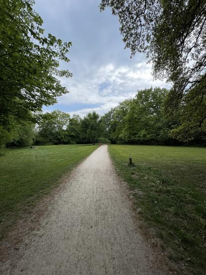 Ein landschaftlich reizvoller Weg, der durch einen üppigen grünen Park führt, der von Bäumen und einem bewölkten Himmel darüber gesänkt wird. Der Weg ist sandig und erstreckt sich in Richtung einer grasbewachsenen Fläche in der Ferne.