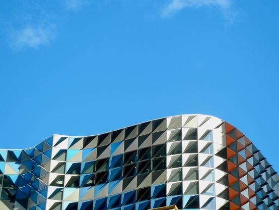 Geometric facade of the RMIT Design Hub building in Melbourne against a clear blue sky, featuring a distinctive curved roofline and triangular panels in white, blue, and orange creating a modern architectural pattern.
