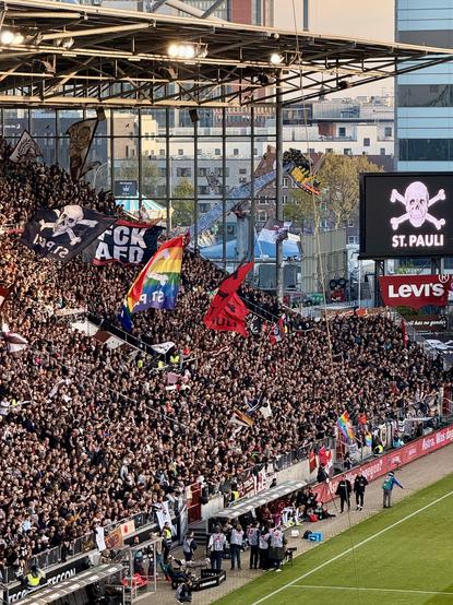 Am Millerntor: Blick auf die vollbesetzte Gegengerade kurz vor dem Beginn des Heimspiels gegen Bayer Leverkusen, das am Ende 1:1 ausging. Zu sehen sind neben dem großen St. Pauli Totenkopf auf der Leinwand diverse große Flaggen im Publikum, wie immer stabil in Regenbogenfarben oder mit eindeutigem #FCKAFD Statement.