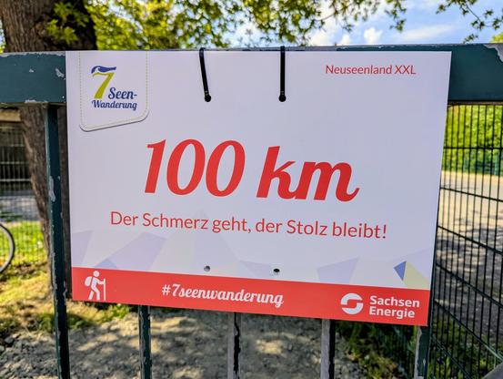 Das Bild zeigt ein Schild der "7-Seen-Wanderung" mit der Aufschrift "100 km". Darüber steht "Neuseenland XXL", darunter der Motivationsspruch: "Der Schmerz geht, der Stolz bleibt!" Unten ist das Logo von "Sachsen Energie" sowie der Hashtag "#7seenwanderung" zu sehen. Das Schild ist an einem Metallzaun befestigt.