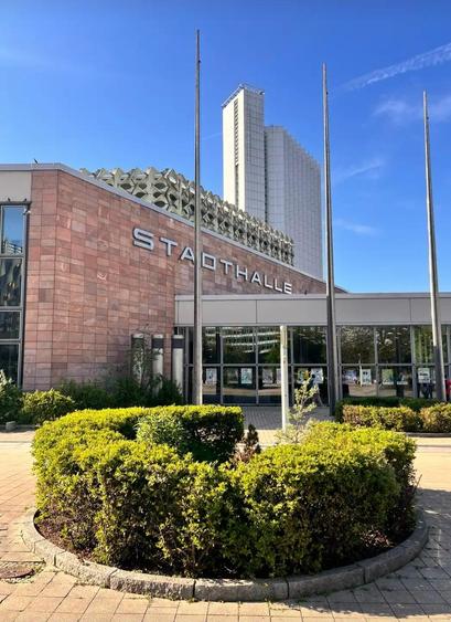 Blick auf die Stadthalle Chemnitz. Im Vordergrund eine kreisrunde Hecke, dahinter ein rosteoter Baukörper, an den eine Glasfassade anschließt. Im Hintergrund ein Hochhaus