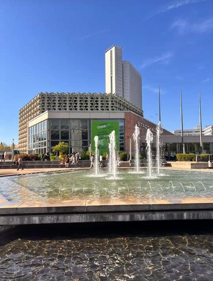 Blick auf die Stadthalle Chemnitz. Im Vordergrund ein Springbrunnen, dahinter die Stadthalle mit der Betonsteinfassade, im Hintergrund ein Hochhaus