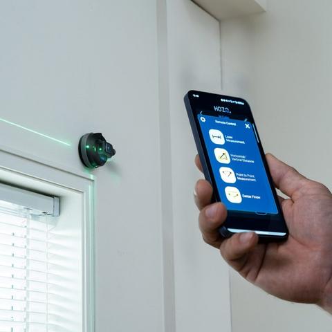 The image shows a close-up of a hand holding a smartphone displaying an app interface. The app is titled "HOZO" and offers several features: "Remote Control," "Laser Measurement," "Horizontal/Vertical Distance," "Point to Point Measurement," and "Center Finder." The smartphone is held in front of a wall-mounted device emitting a green laser line, which is used for measuring distances. The device is circular with a black body and green lights, mounted on a white wall near a window with partially open blinds. The laser line is visible on the wall, indicating the device's function. The smartphone screen is blue with white text and icons, and the time displayed is 12:49.