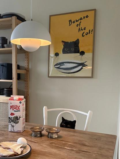 poster of a black cat peering over the edge of a table at a plate with a fish on it hanging over a very similar looking black cat peering over the edge of a dinner table at a plate of bread, cheese & an egg