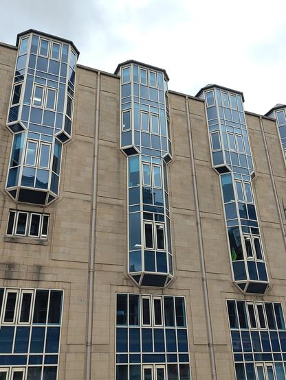 A portrait view of 6 art deco style compound windows in a modern sandstone clad building. The glass has a blue sheen, the frames white. There are three bay windows that break the roofline and of varying height, shortest on left. Below three large flat Georgian panel fixed lights