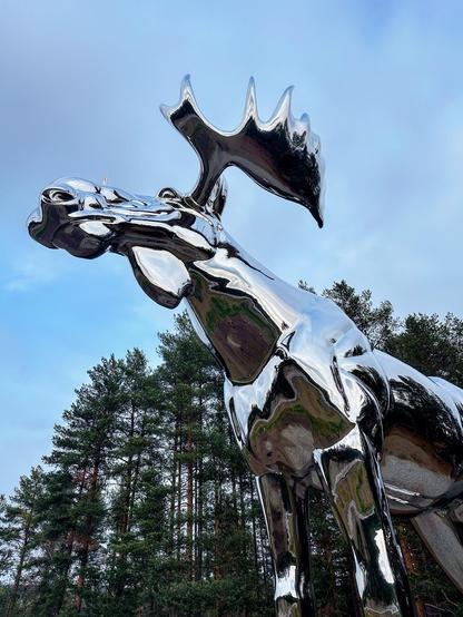 This image features a large, shiny, metallic sculpture of an abstract moose. The sculpture is highly reflective, almost like a mirror, and captures the surrounding environment on its surface, including trees and the sky. The background shows a wooded area with tall pine trees under a cloudy sky. The sculpture's exaggerated and stylized features, especially the antlers, add to its modern and artistic appearance.