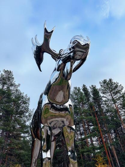 This image shows a shiny, metallic sculpture of what looks like a stylized deer, set against a backdrop of pine trees and a blue sky with light clouds. The sculpture has a particularly abstract and modern design, with smooth, reflective surfaces that mirror the surrounding environment. Its antlers are prominently displayed and exaggerated in size relative to its head and body, enhancing its artistic and dramatic effect. The sculpture is located outdoors, likely in a park or a public art installation area.