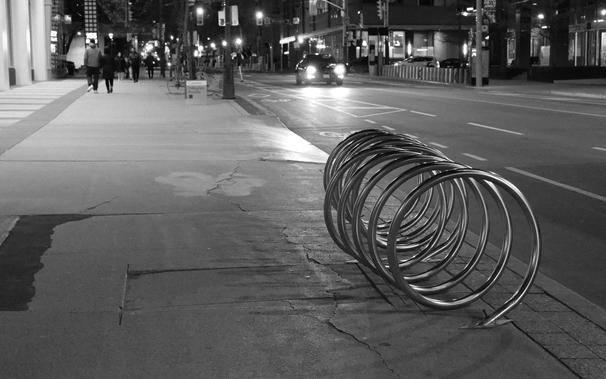 Night, In the foreground, just right of centre, is a bicycle rack, shaped like a coiled spring laid on its side. Its surface is shiny, reflecting the lights. In a band at the top of the image are the background lights of buildings and traffic, along with the small figures of pedestrians walking toward the camera.