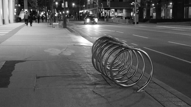 Night, In the foreground, just right of centre, is a bicycle rack, shaped like a coiled spring laid on its side. Its surface is shiny, reflecting the lights. In a band at the top of the image are the background lights of buildings and traffic, along with the small figures of pedestrians walking toward the camera.