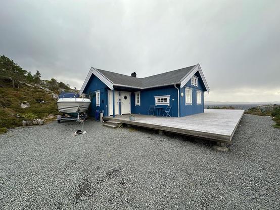 The image shows a charming blue house with white trimmings and a substantial wooden deck area. Beside the house, there is a boat on a trailer, indicating proximity to a water body or the homeowner's interest in boating. The setting appears remote with a natural, rocky landscape surrounding the house, and a cloudy sky suggests overcast weather conditions. Lawn chairs on the deck imply a place for relaxation and enjoyment of the outdoor environment.