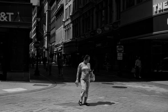 Black and white photo of a broad pedestrian intersection, with buildings emerging from shadows in the background. A lone woman walks through the center of the intersection, glancing at her phone.