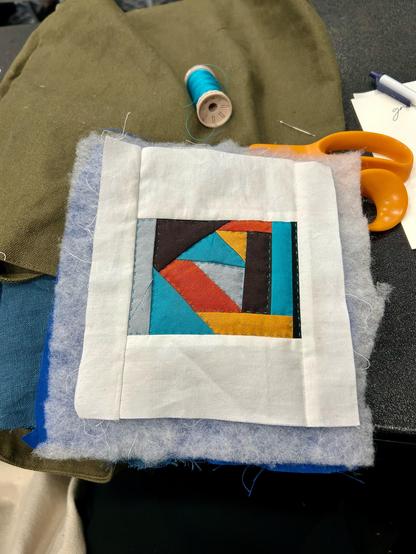 A mini quilt with turquoise, orange, yellow, brown, black, and gray fabric in a central motif with white borders. The  central part involves triangles in a kind of nesting with some slightly uneven rectangles on the sides.