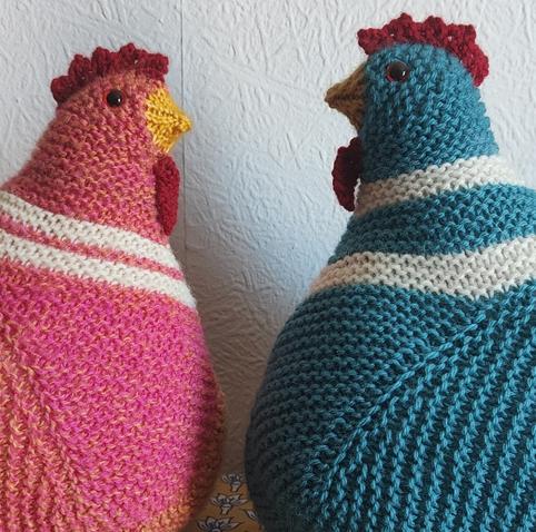 Two knitted chickens - one pink and orange, and one a teal-ish colour - seen sideways on and facing each other against a white walk.