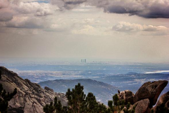 A wide shot captures a rugged, rocky mountain range in the foreground, dotted with evergreen trees. In the distance, a hazy valley unfolds with hints of the Madrid skyline and a body of water under a cloudy sky. A lone vulture sits on a rock near the bottom of the image.