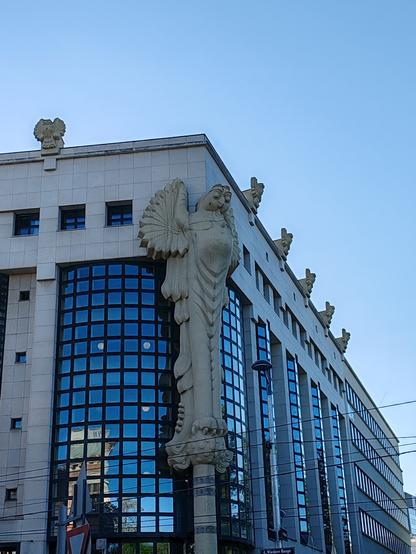 Picture of the corner of the outside of a modern building. The sky above is cloudless and blue. The corner is adorned with what looks like an Egyptian style representation of a bird standing on a pillar, and extends from the ground nearly up to the top of the building. Along the top edges, there are smaller owl-like sculptures which serve as gargoyles. The windows of the building are delimited by black metal square frames, with the corner window extending along two sides from the corner pillar statue.