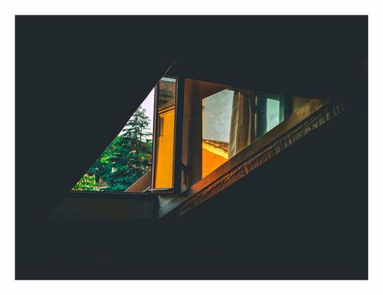 A photo of a window and mirror at sunset. They are positioned it such a way that they appear to be two triangles stuck together creating a rhombus like shape. Outside the window you can see some green trees and a bit of sky. The mirror reflects some yellow buildings. The rest of the photo is black and highlights the weird shape of the objects.