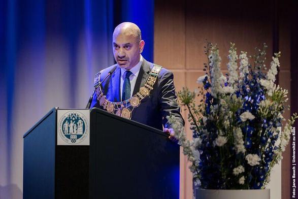 Mann im Anzug am Rednerpult, trägt eine goldene Amtskette mit Medaillons. Das Podium zeigt ein Emblem, daneben ein Blumenarrangement.