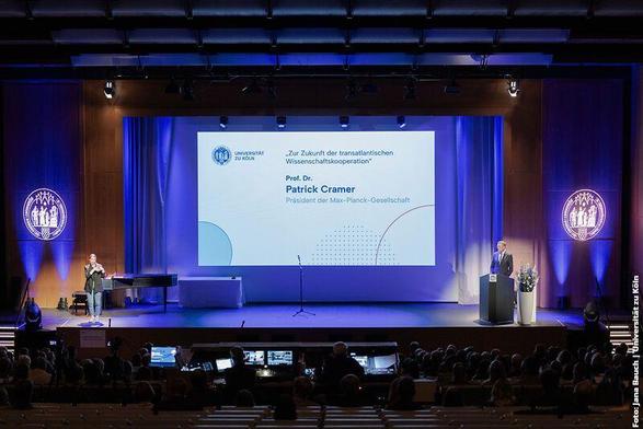 Aula der Universität zu Köln, eine große Leinwand zeigt das Thema 'Zur Zukunft der transatlantischen Wissenschaftskooperation', Prof. Dr. Patrick Cramer spricht als Präsident der Max-Planck-Gesellschaft. Links ein Flügel, rechts ein Rednerpult mit Blumen.