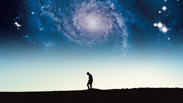 Im unteren Bildraum erkennt man mittig die Silhouette einer Person, den Blick auf den steinigen Boden unter sich gerichtet. Über ihr erstrahlt eine Galaxie im blauen Abendhimmel.