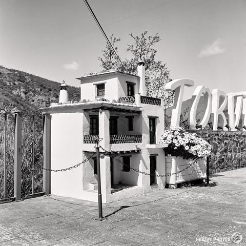A black and white photograph captures a brick pathway in Torvizcón with a scale model of one of the village houses. A large planter with flowers stands next to it. In the background, you can see a mountain landscape, while a sign with partially visible letters sof the village name tands out against the clear sky.