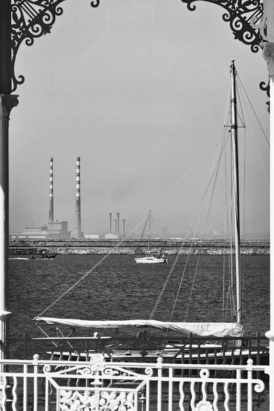 A black and white photograph taken from a decorative archway, showing a calm harbor with sailboats, distant industrial chimneys, and the coastline.