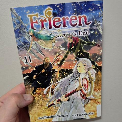 Photograph of a white person's hand holding Frieren: Beyond Journey's End, volume 11. Image shows the titular Frieren centralized ready to strike with her staff at the ready with various characters behind her battling including an older, bearded gentleman (Denken) and a few demons. The lower half of the scene is covered in golden layer. The upper half is the night sky, largely.