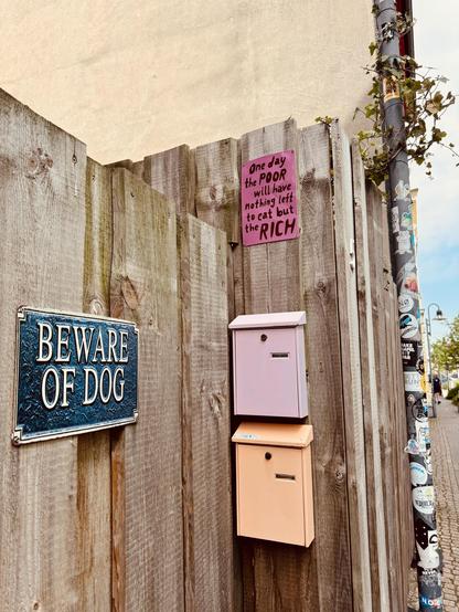 Das Bild zeigt einen Holzzaun mit einem Schild "Beware of Dog", zwei Briefkästen und einem handgeschriebenen Zettel mit der Botschaft "One day the poor will have nothing left to eat but the rich", während im Hintergrund ein Laternenpfahl mit Plakaten zu sehen ist.