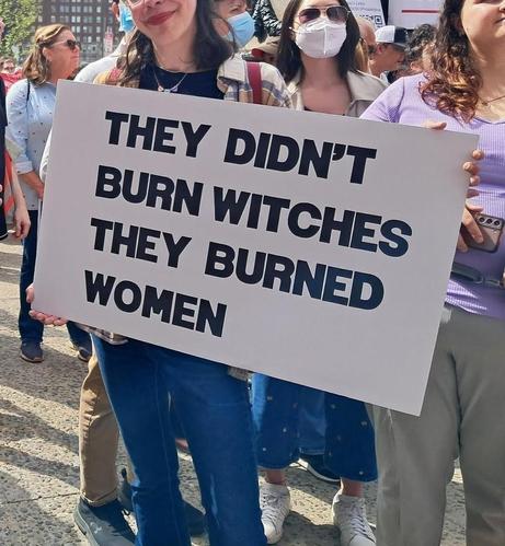 A woman holds a protest sign on a street in Philadelphia, as she stands with other protesters.  The sign reads, "They didn't burn witches, they burned women."