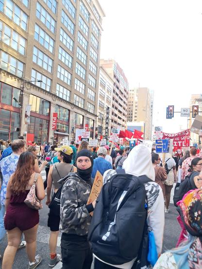 A huge crowd of diverse people are walking together in the streets of Philadelphia, holding protest signs.