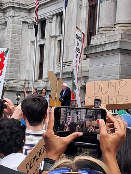 Senator Bernie Sanders is standing above the crowd of protestors in Philadelphia.  Many in the crowd are photographing him as he addresses them.  A protest sign can be seen which reads, "Dump Trump."