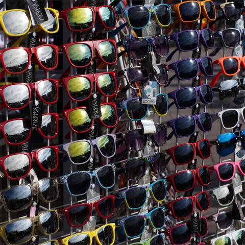 A display sunglasses with colourful frames, arranged in neat rows on a rack outdoors. Sunlight is reflecting from them.