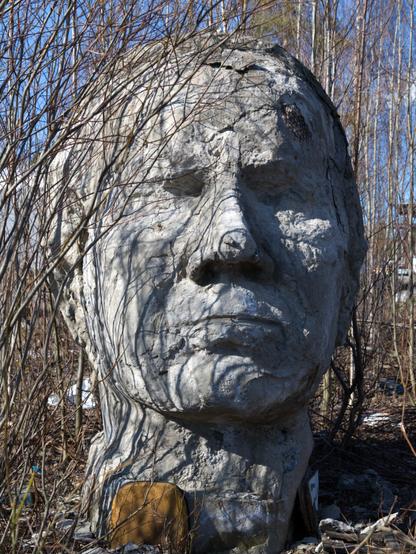 A massive, somewhat weathered concrete statue head, amidst bare branches in springtime, under blue sky. Ground is full of dead leaves, with some debris lying about.