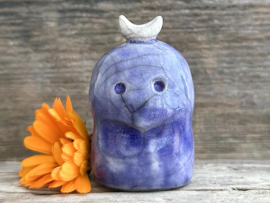 against a background of grey-brown driftwood, a raku ceramic kodama glazed in blue. it has wide eyes, a slightly sad and worried faced, and a white crescent moon on its head. 
there's an orange calendula flower next to it on the left.