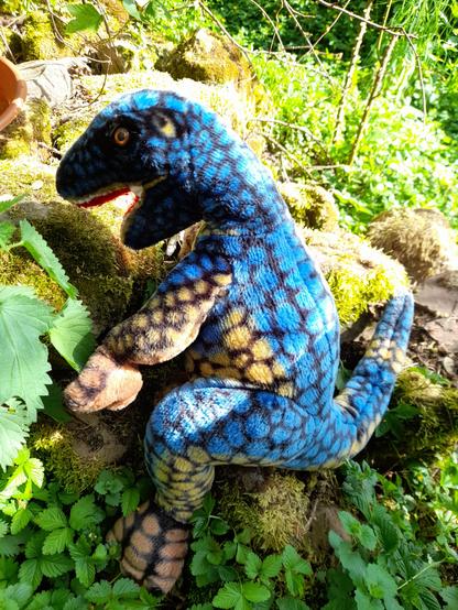 A rather big lezard-headed blue plush dinosaur climbin on some sandstones between wild herbs.