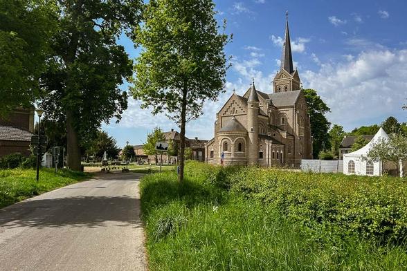 Das Bild zeigt eine idyllische Landschaft mit einem imposanten Kirchturm im Hintergrund. Die Kirche, mit ihrem hohen, schmalen Turm und gotischen Elementen, ist aus Stein gebaut und hat mehrere Türme und Fenster mit Bogen. Sie steht auf einem gepflegten Rasen, der von üppigem Grün umgeben ist. Ein asphaltierter Weg führt durch das Bild, beginnend im Vordergrund und führt in Richtung Kirche. Links und rechts des Weges stehen hohe Bäume, die Schatten werfen. Im Hintergrund sind weitere Gebäude und ein weißer Zeltstand zu sehen. Der Himmel ist blau mit einigen weißen Wolken, was auf einen sonnigen Tag hinweist.