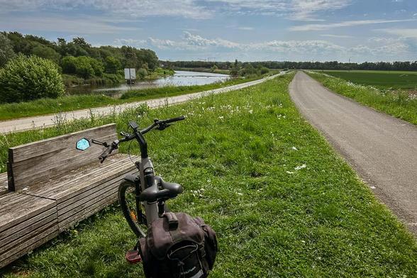 as Bild zeigt eine ruhige Landschaft mit einem Fahrrad, das an einem Holzbankplatz lehnt. Der Weg, auf dem das Fahrrad steht, führt entlang eines Kanals und ist von grünem Gras und wilden Blumen umgeben. Im Hintergrund erstreckt sich ein Feld, das von Bäumen gesäumt wird. Der Himmel ist blau mit einigen weißen Wolken. Ein Schild am Kanalrand ist sichtbar, aber keine Texte sind lesbar. Die Szene vermittelt ein Gefühl von Frieden und Natur.