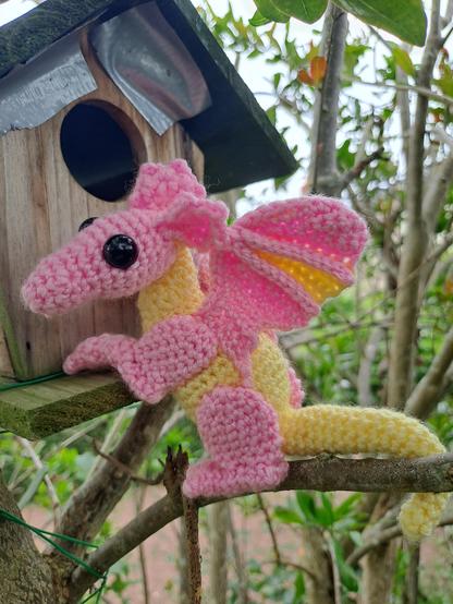 Petit dragon au crochet debout assis sur ses pattes arrière : corps jaune, pattes rose, ailes bicolores, tête rose. 
Il a l'air déçu que la cabane soit vide :'(