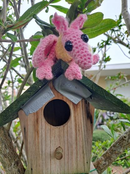 Petit dragon au crochet debout assis sur ses pattes arrière : corps jaune, pattes rose, ailes bicolores, tête rose. Il est désormais perché sur la cabane.