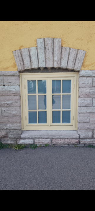 A window at ground level, in a yellow building where the bottom of the building is grey bricks and there is also a few grey bricks above the window. The wooden-frame directly around the window is yellow.