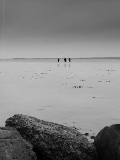 The image shows a black and white scene of three people standing in shallow water, possibly engaged in fishing, as they are holding fishing rods. In the foreground, there are large rocks, and the background features a distant horizon and a calm, expansive body of water. The overall atmosphere is serene and minimalistic.