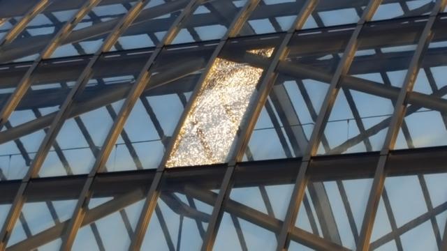 A foto of of both the front and back of an arched roof consisting of rows of portrait oriented, steel framed, safety glass panes. The middle pane in the front has splintered and is refractie the sunlight, giving it a golden sheen and making it stand out from the grey girders and blue sky background.
