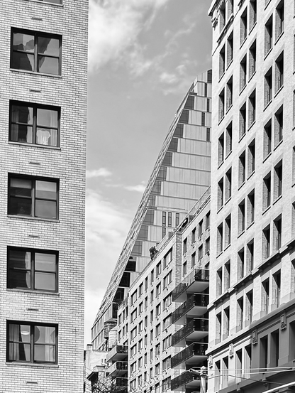Black and white photo of a close-up cityscape. On the left is a 1960s style white brick building and on the right is a more simple stone building with some brick buildings next to it. Further down the street is a glass building that goes up about nine stories and then slopesto the right making it look like a pyramid of glass windows