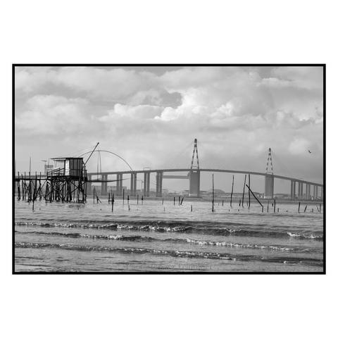 (English follows) Photographie noir et blanc, format paysage, ratio 3x2. La plus grande partie de l'image est occupée par le lit de la Loire (premier plan) et le ciel. Vers le milieu de l'image, se découpe une pêcherie s'avançant dans les eaux du fleuve. Le pont de Saint-Nazaire traverse toute l'image, comme dans le prolongement de la pêcherie. L'image offre un contraste entre les éléments naturels et industriels de l'estuaire de la Loire. 

Black and white photograph, landscape format, 3x2 ratio. Most of the image is taken up by the bed of the Loire (foreground) and the sky. Towards the middle of the image, a fishery juts out into the river. The Saint-Nazaire bridge crosses the entire image, as if an extension of the fishery. The image contrasts the natural and industrial elements of the Loire estuary.