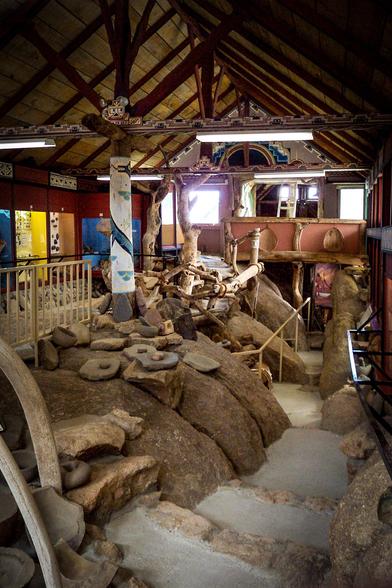 The image depicts an indoor archaeological exhibit with a rustic and historical ambiance. The space features a high, wooden ceiling with exposed beams and a decorative pattern along the edges. The walls are adorned with colorful, geometric patterns, enhancing the cultural theme. The floor is uneven, with large rocks and stones scattered throughout, creating a naturalistic setting. Various artifacts, including stone tools and pottery, are displayed on the ground, some partially buried, suggesting an excavation site. A large, carved wooden wheel is visible on the left side, adding to the historical context. The exhibit includes a raised platform with a railing, providing a vantage point to view the artifacts. Natural light filters in through windows, illuminating the space and highlighting the textures and colors of the exhibits. The overall layout and design suggest an immersive experience, allowing visitors to explore and learn about the historical context of the artifacts.

Provided by @altbot, generated privately and locally using Ovis2-8B