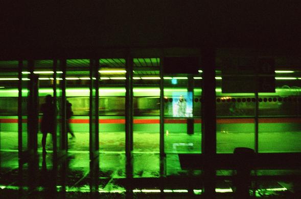 A long exposure photograph of a metro station behind glass wall with train passing and some people standing on the platform. The scene has strong green hue.
