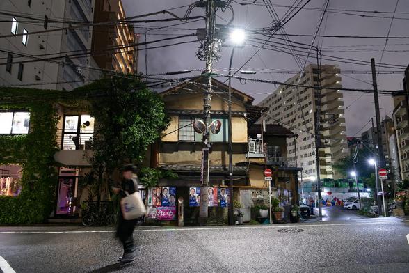 Urban night scene featuring a mix of traditional and modern buildings. A person walks along a wet street, with greenery on one side and a towering apartment complex in the background. Streetlights illuminate the area, revealing advertisements and traffic signs.