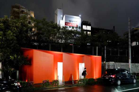 A bright red structure with multiple white doors is illuminated at night, set against a dark sky. Nearby, a person walks past the building, while a black taxi is parked on the side of the road. Lush greenery and plants are visible.